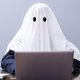 A person wearing a white sheet as a ghost costume types on a computer.